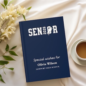 May include: A navy blue hardcover journal with the text "SENIOR 2026" and "Special wishes for Olivia Wilson, Newport High School" in white. The journal is next to a cup of coffee and white flowers.