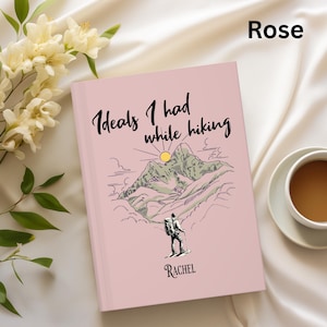 May include: A pink journal with the text "Ideals I had while hiking" and a mountain illustration. The journal has a hiker illustration and the name "Rachel" at the bottom. A cup of coffee and flowers are next to the journal.