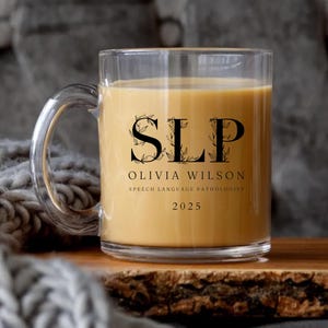 May include: A clear glass mug filled with a light brown liquid. The mug features the letters "SLP" with floral accents, the name "OLIVIA WILSON", and the text "SPEECH LANGUAGE PATHOLOGIST" and "2025".