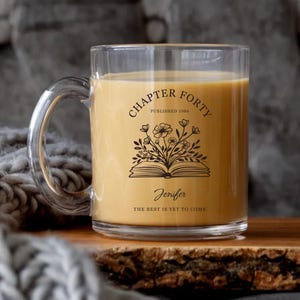 May include: A clear glass mug filled with a light-colored beverage. The mug features a black floral design with the text "CHAPTER FORTY PUBLISHED 1986" and the name "Jenifer" with the phrase "THE BEST IS YET TO COME".