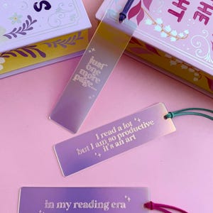 May include: Three iridescent bookmarks with different quotes. The first bookmark says "just one more page". The second bookmark says "I read a lot ++ but I am so productive it's an art". The third bookmark says "in my reading era".