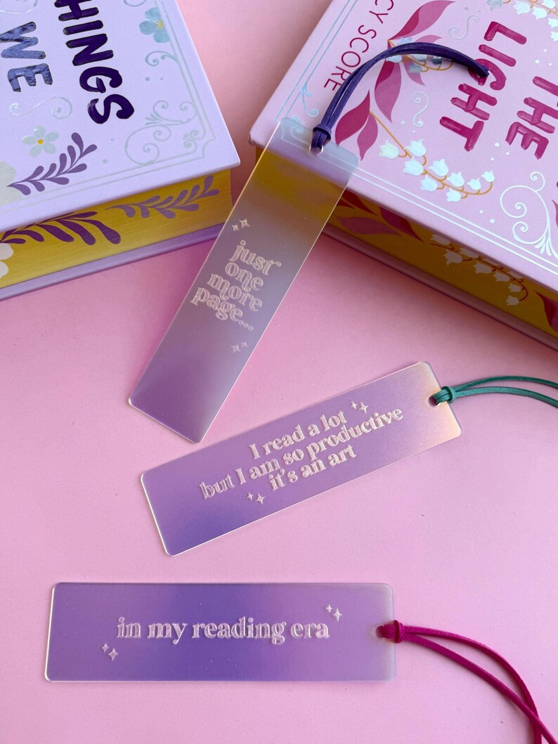 May include: Three iridescent bookmarks with different quotes. The first bookmark says "just one more page". The second bookmark says "I read a lot ++ but I am so productive it's an art". The third bookmark says "in my reading era".
