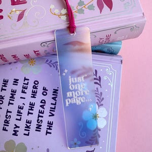 May include: A holographic bookmark with a blue and purple gradient background. The bookmark features a white floral design and the text "just one more page..." in a white, cursive font.