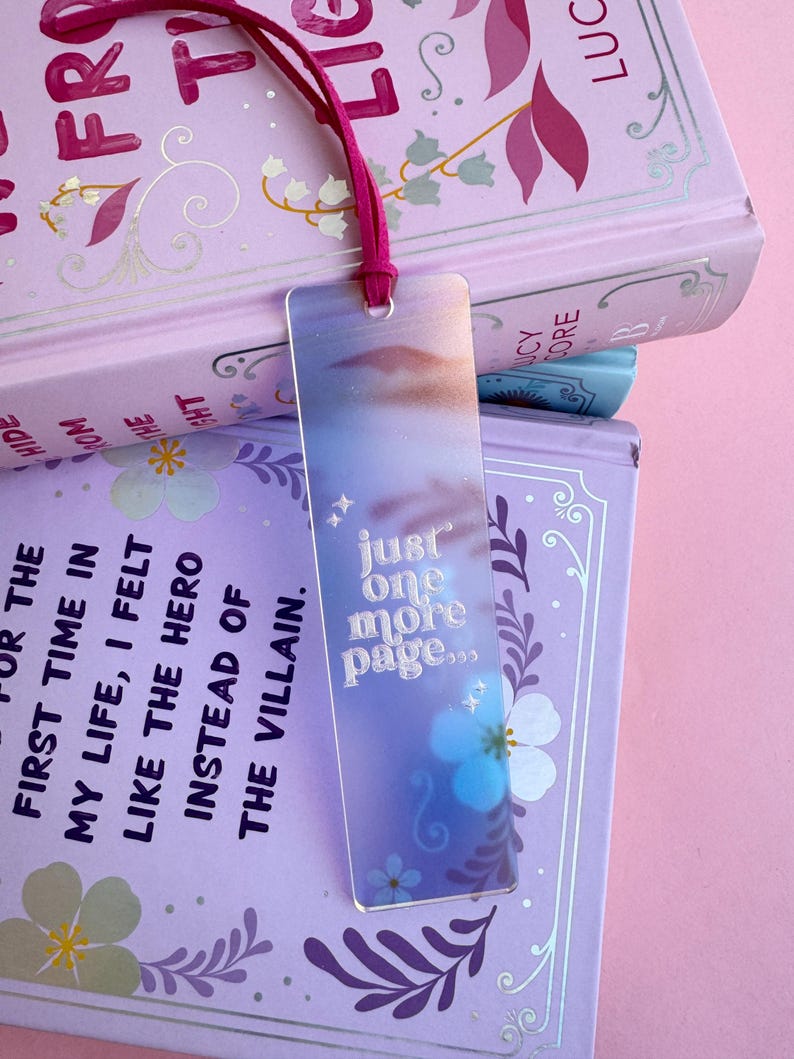 May include: A holographic bookmark with a blue and purple gradient background. The bookmark features a white floral design and the text "just one more page..." in a white, cursive font.