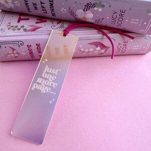 May include: A pink and iridescent bookmark with the text "just one more page..." in white. The bookmark is laying on top of two books with floral designs and the text "Things We Left Behind" and "Lucy Score" on the covers.