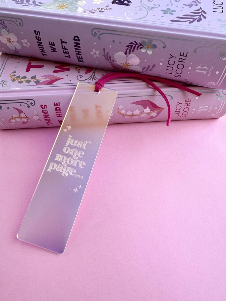 May include: A pink and iridescent bookmark with the text "just one more page..." in white. The bookmark is laying on top of two books with floral designs and the text "Things We Left Behind" and "Lucy Score" on the covers.