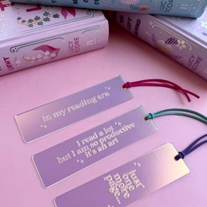 May include: Three iridescent bookmarks with pink, purple, and blue hues. The bookmarks feature quotes about reading: "in my reading era", "I read a lot but I am so productive it's an art", and "just one more page...".
