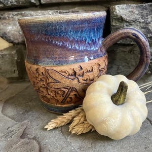 May include: A ceramic mug with a blue and purple glazed upper section and a textured tan lower section featuring a dragonfly relief. A small white decorative pumpkin and dried wheat stalks are positioned in front of the mug.