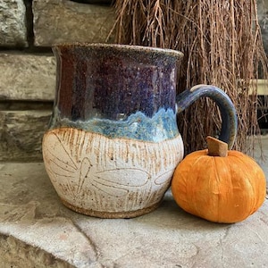 May include: A handmade ceramic mug with a blue and brown glaze, featuring a dragonfly design. An orange pumpkin sits next to the mug. The mug has a curved handle and a wide base. The mug is approximately 4 inches tall.