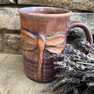 May include: A ceramic mug with a glazed finish in shades of brown, purple, and blue, featuring a raised, sculpted dragonfly design on the front. The mug has a curved handle and a smooth rim, showcasing a blend of earthy and cool-toned colors.