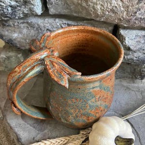 May include: A ceramic mug with a mottled orange and teal glaze featuring a sculpted dragonfly on the handle and a floral pattern embossed on the body. The mug is accompanied by a small white decorative pumpkin and dried wheat stalks.