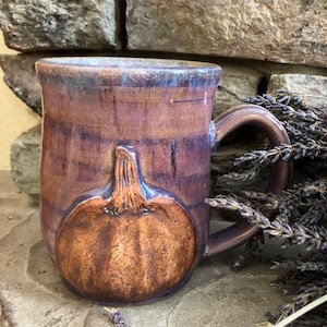 Può includere: Una tazza in ceramica con finitura smaltata nei toni del marrone e del viola, caratterizzata da un rilievo a forma di zucca testurizzato sulla parte anteriore. La tazza presenta un manico robusto e un bordo liscio, dall'aspetto artigianale.