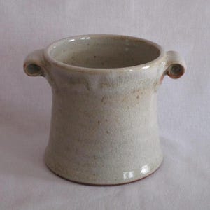 May include: A handmade, off-white ceramic vase with two small, curved handles. The vase has a cylindrical shape with a slightly flared base and a textured surface. The interior is visible, and the rim is smooth. The vase is approximately 5 inches tall.