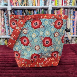 Può includere: Una borsa in tessuto con coulisse su sfondo azzurro, decorata con un motivo di grandi fiori rossi e piccoli fiori gialli. La borsa presenta un bordo superiore rosso e una fascia inferiore rossa con stampa floreale, oltre a un manico rosso.