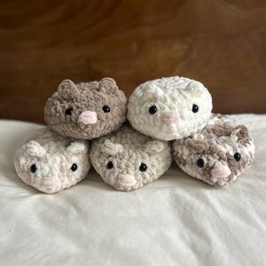 May include: A collection of five handmade crochet animal plushies. The plushies are in various colors, including white, brown, and gray, with pink noses and black eyes. The toys are arranged on a white surface.