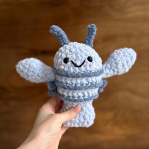 May include: A light blue crochet lobster plush toy with a smiling face, black eyes, and pink cheeks. The lobster has two antennae and two large claws. The body is striped with a darker blue yarn.