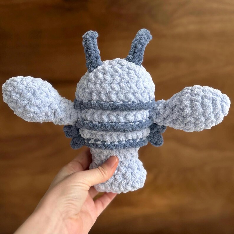 May include: A handmade, light blue crochet lobster plush toy with darker blue accents. The plush has two large claws, two antennae, and a segmented body. The toy is soft and fluffy, perfect for children or as a decorative item.