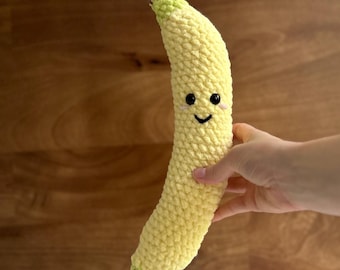 May include: A handmade, smiling banana plush toy in shades of yellow, green, and brown. The banana has a friendly face with black eyes and a curved mouth. The plush is held against a wooden background.