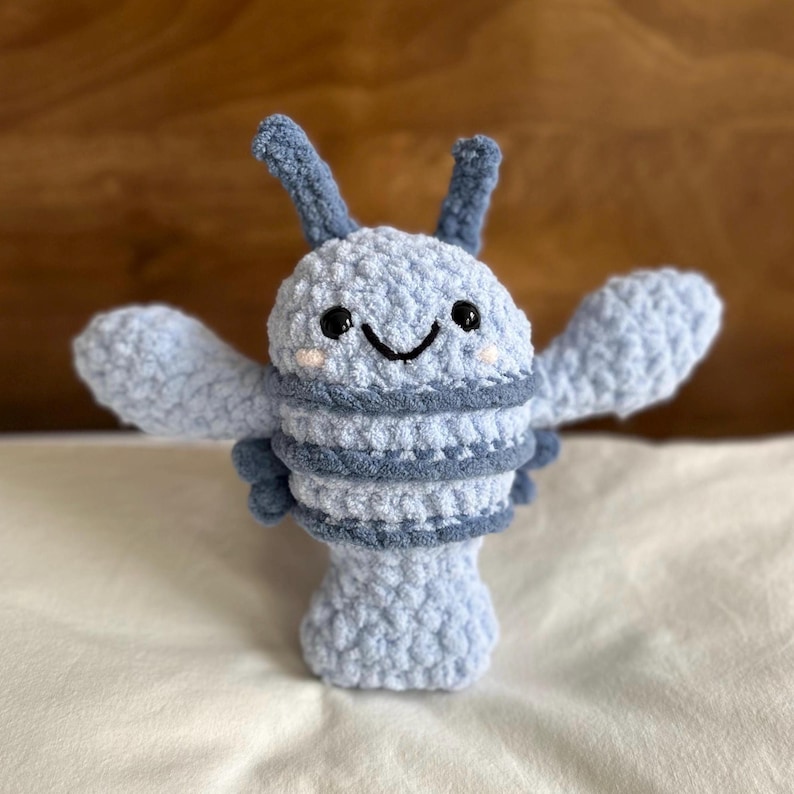 May include: A handmade, light blue crochet creature with dark blue accents. The plush toy has a smiling face, two antennae, and outstretched arms. The body features horizontal stripes and a rounded base, creating a whimsical design.