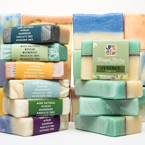 May include: A stack of handmade soap bars in various colors and patterns, including green, blue, yellow, and white. The soap bars are labeled with the text "100% Natural", "Vegan", "Handmade", "Sulfate Free", and "Paraben Free". The top bar is labeled "Fleegal Farma" and "SYNERGY".