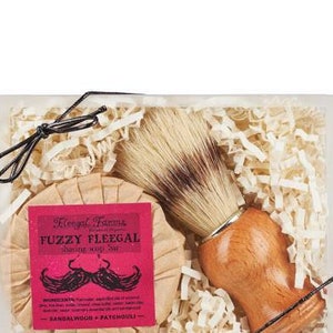 A natural boar bristle shaving brush next to a round Fuzzy Fleegal shave soap bar that builds a foamy lather.