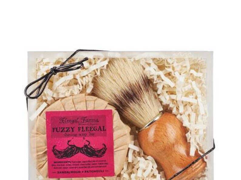 A natural boar bristle shaving brush next to a round Fuzzy Fleegal shave soap bar that builds a foamy lather.