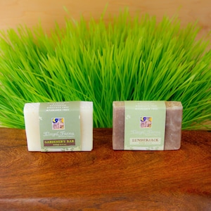 May include: Two bars of soap, one white and one brown, with labels that say "Gardener's Bar" and "Lumberjack". The labels are white with green text and a small image of a moon and flowers. The soap bars are sitting on a wooden surface in front of a bed of green grass.
