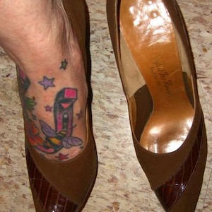 May include: A pair of brown pointed-toe pumps featuring a matte suede finish on the heel and back, contrasted by a glossy, dark brown textured panel across the toe box. The interior lining is a smooth, tan-colored material with visible branding.