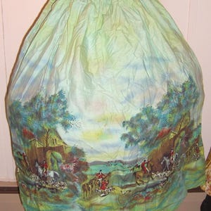 May include: A full-length, gathered skirt in a pale mint green hue, featuring a printed landscape scene along the hem. The print depicts figures on horseback accompanied by hounds in a countryside setting with trees, hills, and stone structures.