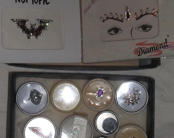 May include: A collection of adhesive face gems and body stickers. The set includes a bat-shaped gem labeled Hot Topic, a Mini Bridal Diamond set with forehead decals, and a black box containing eight small circular containers with various crystal-encrusted bindi-style ornaments in silver, purple, and clear tones.