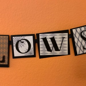 Pu&ograve; includere: Uno striscione di Halloween bianco e nero con la parola "LOWS" in lettere nere su sfondo bianco. Ogni lettera &egrave; su un quadrato separato con un motivo diverso. La "L" &egrave; su un motivo a rombi marrone e bianco, la "O" &egrave; su uno sfondo bianco con una ragnatela nera, la "W" &egrave; su un motivo a rombi bianco e grigio, e la "S" &egrave; su un motivo a quadri bianco e grigio.