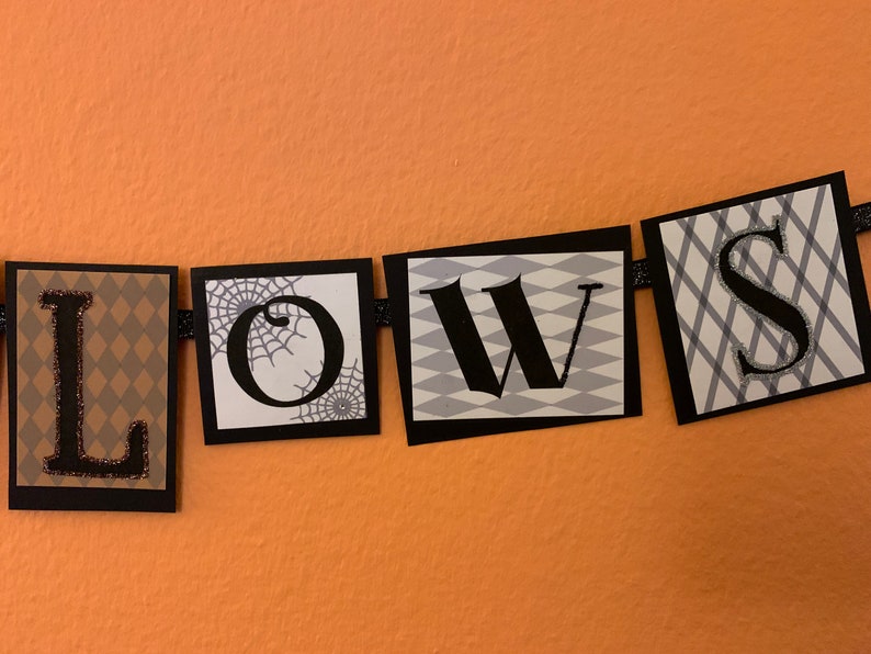 Pu&ograve; includere: Uno striscione di Halloween bianco e nero con la parola "LOWS" in lettere nere su sfondo bianco. Ogni lettera &egrave; su un quadrato separato con un motivo diverso. La "L" &egrave; su un motivo a rombi marrone e bianco, la "O" &egrave; su uno sfondo bianco con una ragnatela nera, la "W" &egrave; su un motivo a rombi bianco e grigio, e la "S" &egrave; su un motivo a quadri bianco e grigio.