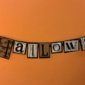Pu&ograve; includere: Uno striscione di Halloween bianco e nero con la parola "Halloweens" scritta in quadrati individuali. Ogni quadrato ha un motivo diverso.
