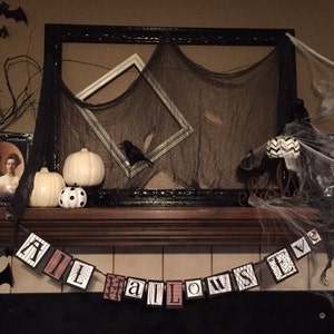 Pu&ograve; includere: Un camino sopra il camino decorato per Halloween con pipistrelli neri, corvi e zucche bianche. Uno striscione bianco e nero con la parola "HALLOWEEN" &egrave; appeso sopra il camino.