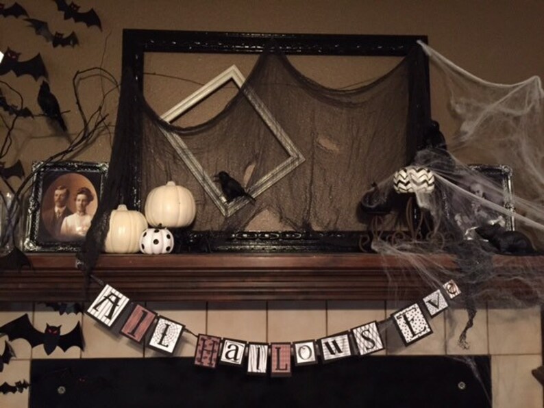 Pu&ograve; includere: Un camino sopra il camino decorato per Halloween con pipistrelli neri, corvi e zucche bianche. Uno striscione bianco e nero con la parola "HALLOWEEN" &egrave; appeso sopra il camino.