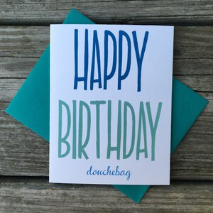 May include: A white card with blue and green lettering that says "Happy Birthday douchebag". The card is sitting on a teal envelope.