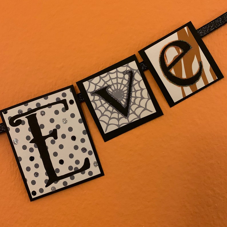 Pu&ograve; includere: Uno striscione bianco e nero con la parola "EVE" scritta in lettere nere. Ogni lettera &egrave; su un foglio di cartoncino separato con un motivo diverso. La "E" &egrave; su uno sfondo bianco con pois neri, la "V" &egrave; su uno sfondo bianco con un disegno di ragnatela nera, e la "E" &egrave; su uno sfondo zebrato marrone e bianco.