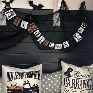 Pu&ograve; includere: Uno striscione di Halloween bianco e nero con la parola "Halloween" scritta in blocchi individuali. Lo striscione &egrave; appeso a un tessuto a rete nero. Due pipistrelli di carta neri sono appesi al soffitto sopra lo striscione. Due cuscini con motivi a tema Halloween sono su un divano grigio. Un cuscino ha il testo "Old Crow Pumpkins" e un camion nero con zucche nel cassone. L'altro cuscino ha il testo "Broom Parking Witches Only 25c All Others Will Be Toad".
