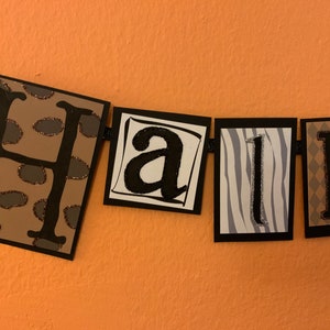 Pu&ograve; includere: Uno striscione bianco e nero con la parola "Hall" scritta in lettere nere con accenti di glitter. Ogni lettera &egrave; su un quadrato separato con uno sfondo di stampa animale diverso. Le lettere sono su uno sfondo marrone.