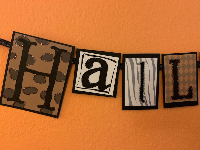 Pu&ograve; includere: Uno striscione bianco e nero con la parola "Hall" scritta in lettere nere con accenti di glitter. Ogni lettera &egrave; su un quadrato separato con uno sfondo di stampa animale diverso. Le lettere sono su uno sfondo marrone.