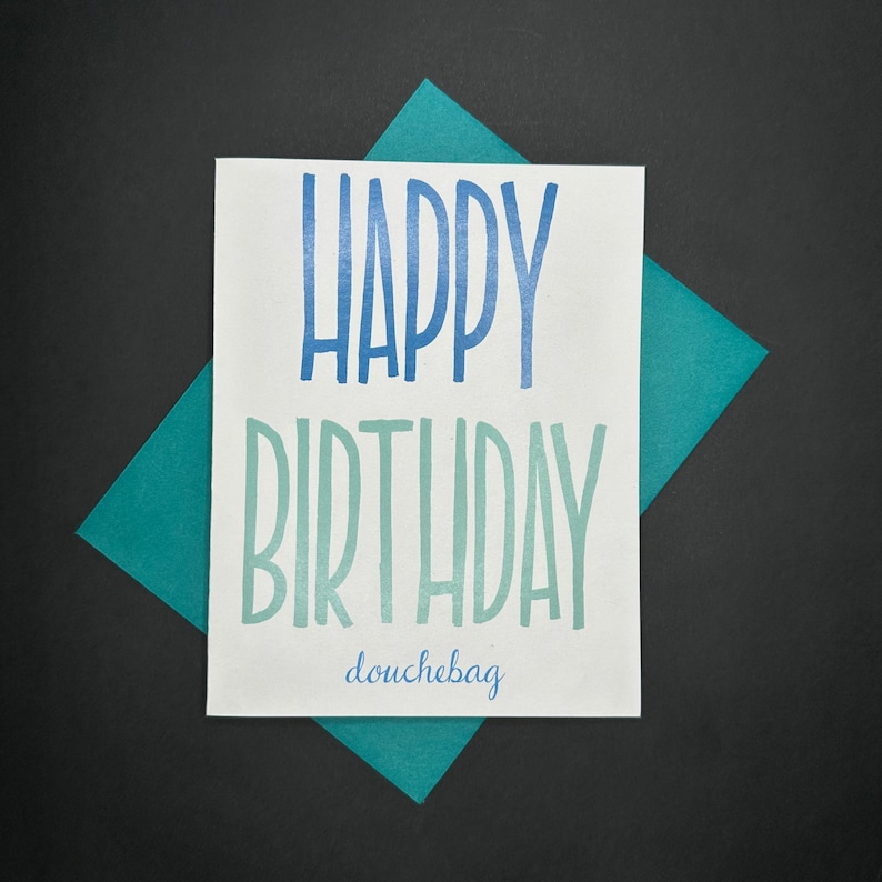 May include: A white card with blue and green text that says "Happy Birthday douchebag". The card is on a teal blue background.