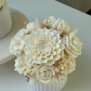 May include: A white, floral-shaped candle arrangement in a small, ribbed, white ceramic pot. The arrangement includes a large dahlia, roses, and other flower shapes, with dried accents. The candle is a decorative home decor item.