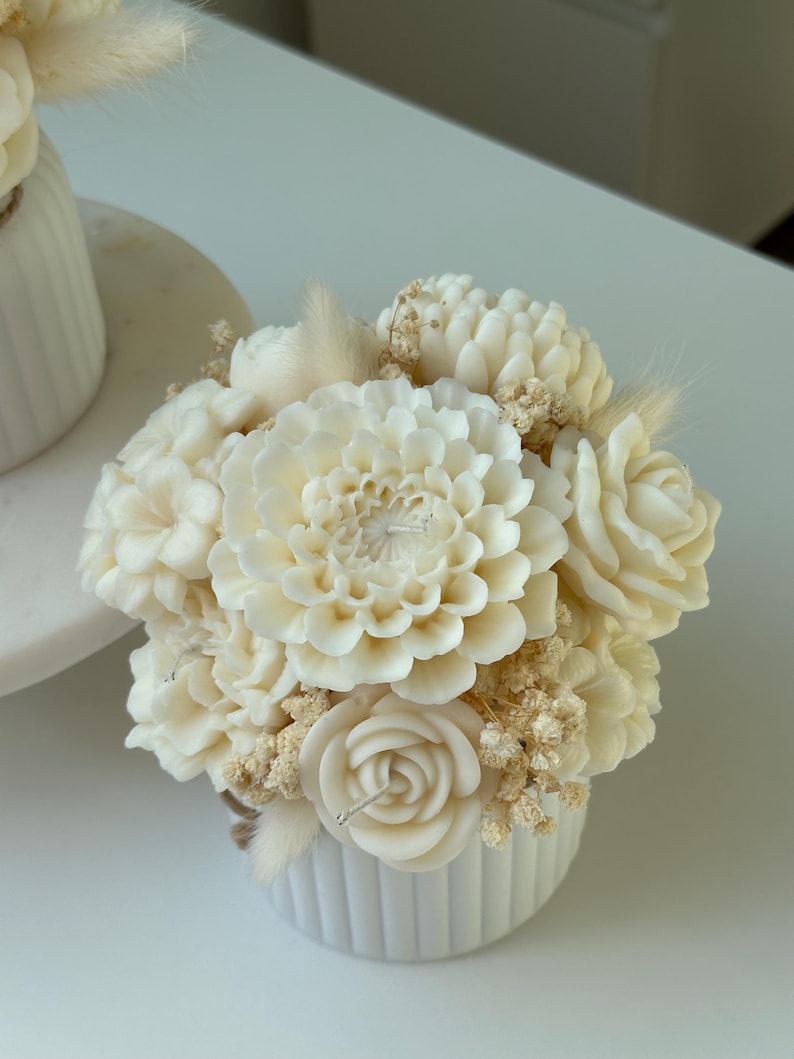 May include: A white, floral-shaped candle arrangement in a small, ribbed, white ceramic pot. The arrangement includes a large dahlia, roses, and other flower shapes, with dried accents. The candle is a decorative home decor item.