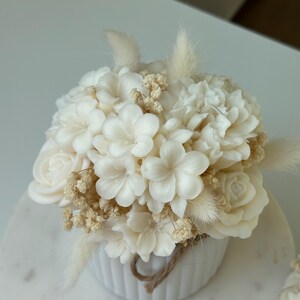 May include: A white floral arrangement in a small, white, ribbed ceramic pot. The arrangement features various white flowers, including roses and other blossoms, with dried accents and a twine detail. The arrangement is on a white marble surface.
