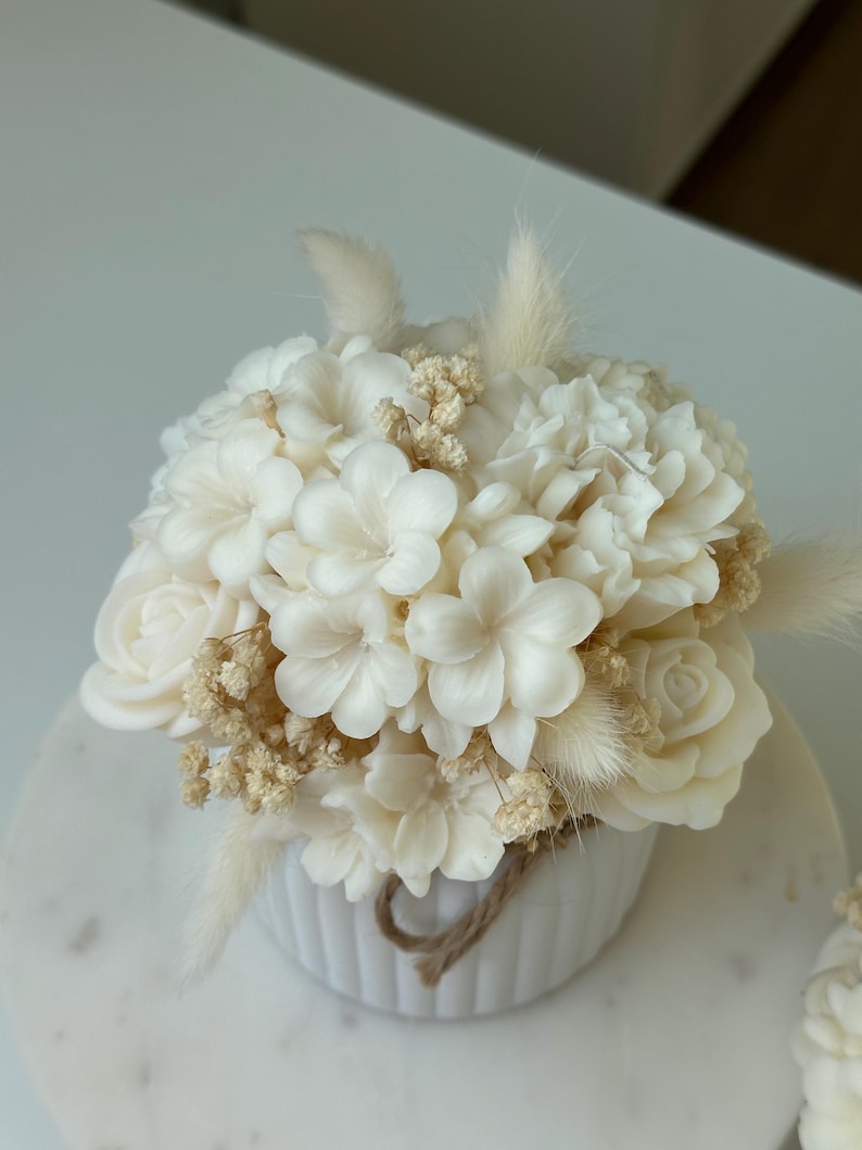 May include: A white floral arrangement in a small, white, ribbed ceramic pot. The arrangement features various white flowers, including roses and other blossoms, with dried accents and a twine detail. The arrangement is on a white marble surface.