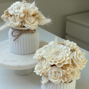 May include: Two white ceramic vases with vertical grooves, each holding a bouquet of ivory-colored flowers. The bouquets include roses, dahlias, and other floral elements, accented with dried grasses. A twine bow adorns one vase.