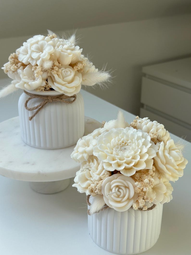 May include: Two white ceramic vases with vertical grooves, each holding a bouquet of ivory-colored flowers. The bouquets include roses, dahlias, and other floral elements, accented with dried grasses. A twine bow adorns one vase.