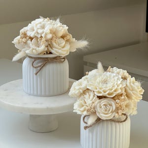 May include: Two white, ribbed ceramic vases, each adorned with a bouquet of cream-colored flowers and dried accents. A twine bow encircles each vase. One vase sits on a white marble cake stand, the other on a white surface.