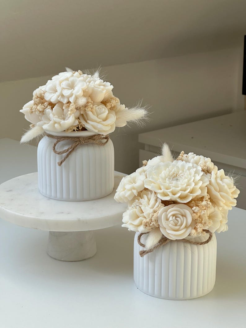 May include: Two white, ribbed ceramic vases, each adorned with a bouquet of cream-colored flowers and dried accents. A twine bow encircles each vase. One vase sits on a white marble cake stand, the other on a white surface.