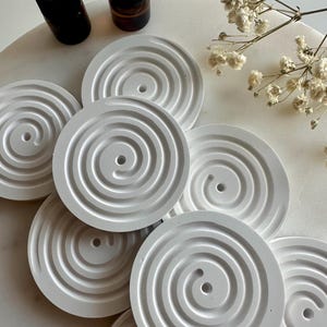 Könnte beinhalten: Weiße Keramik-Untersetzer mit einem Spiraldesign. Die Untersetzer sind in einem Kreis auf einer weißen Oberfläche angeordnet. Zwei kleine Flaschen sind im Hintergrund sichtbar.